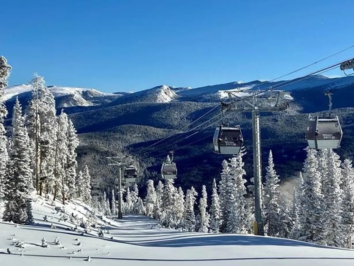 A snowy, mountainous landscape featuring a gondola lift ascending over evergreen trees, with skiers and snowboarders visible below. The dramatic mountain vistas and lift infrastructure set a scene of high-altitude excitement and access to world-class ski terrain.
