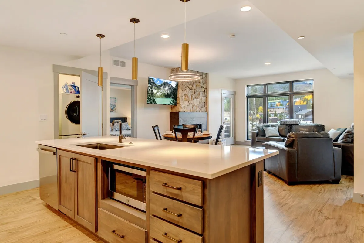 A wide-angle view capturing an open-concept living area featuring a kitchen island, dining table, lounge area with a couch, a flat-screen television, and a stone fireplace. The layout emphasizes flow between cooking, dining, and relaxing spaces, ideal for gatherings.