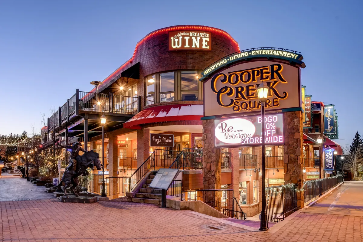 A lively scene of Cooper Creek Square, with its prominent sign, inviting wine shop entrance, outdoor seating, decorative lighting, and a foreground sculpture. The area is bustling with activity, hinting at upscale dining, boutique shopping, and local art—all integral parts of the downtown Winter Park experience.