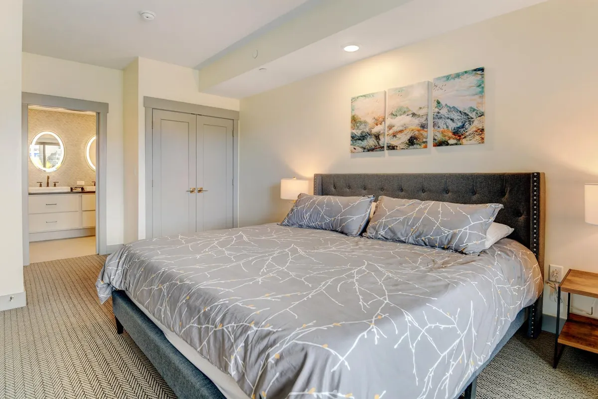 A serene master bedroom featuring a large bed adorned with decorative bedding, soft lighting, and an entrance to an ensuite bathroom. Artwork on the wall and natural light filtering through the windows create a calming, retreat-like atmosphere.