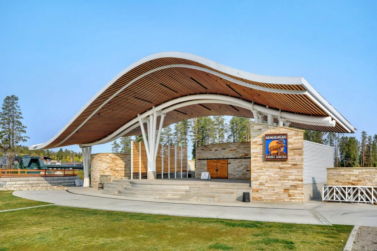 The Rendezvous Events Center's distinctive curved roof and stone facade set the stage for festive outdoor concerts and community events. The shot includes the open stage area, suggesting the excitement of live performances just across the street from the condo.