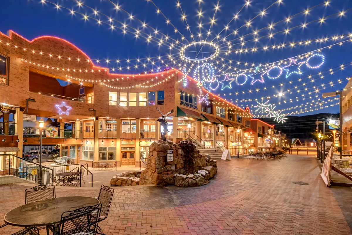 A lively exterior setting featuring decorative string lights, a stone fountain, outdoor seating areas, and attractive building architecture. The scene is bustling and festive, suggesting a space ideal for social gatherings, community events, or evening outings.