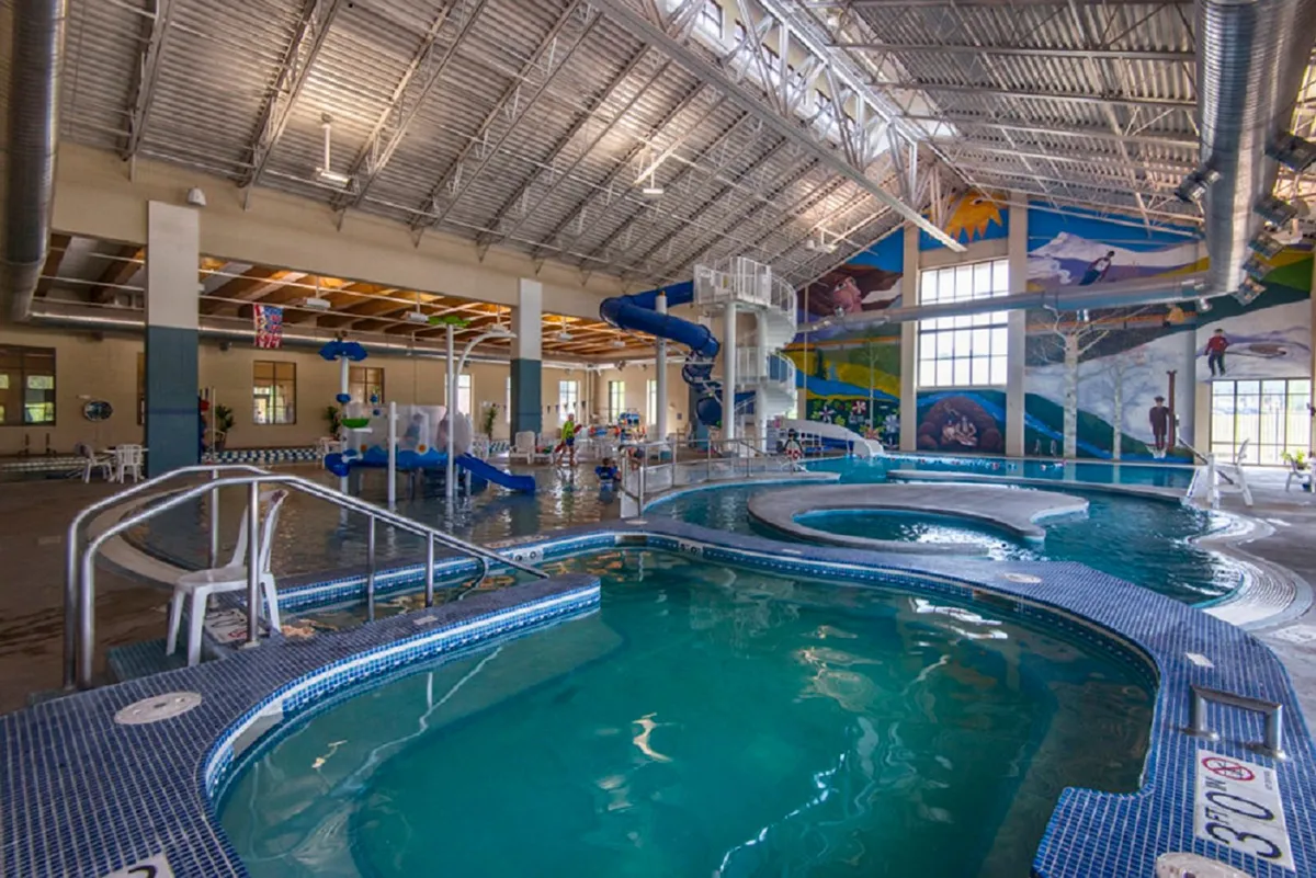 An inviting indoor recreation area with a large swimming pool, colorful water slides, playful murals, and families enjoying themselves in and around the pool. Children are splashing in the water, and several lounge chairs provide space for relaxation.