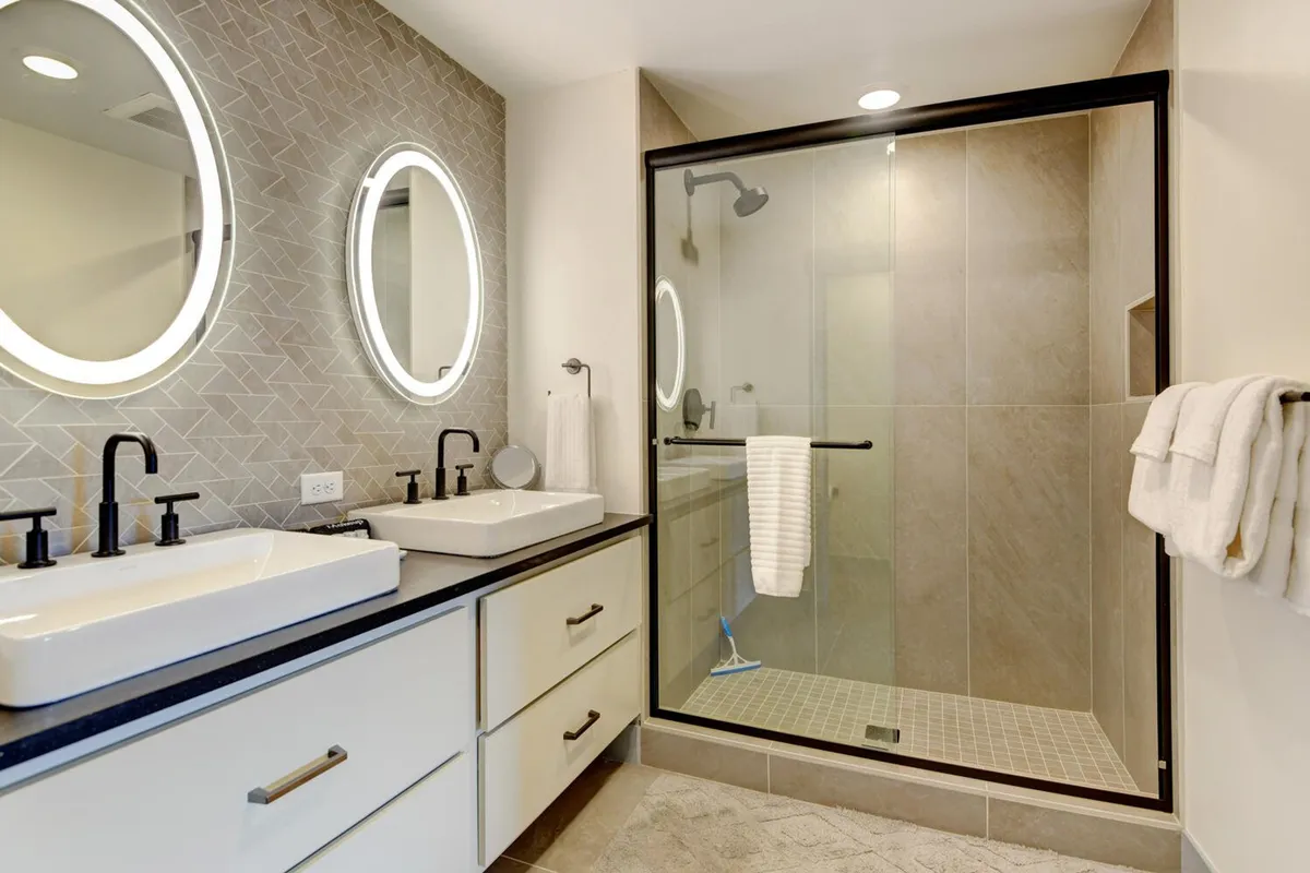 A sophisticated bathroom with double sinks, bright mirrored lighting, and a glass-enclosed shower. The space is clean, modern, and inviting, ideal for refreshing and unwinding.