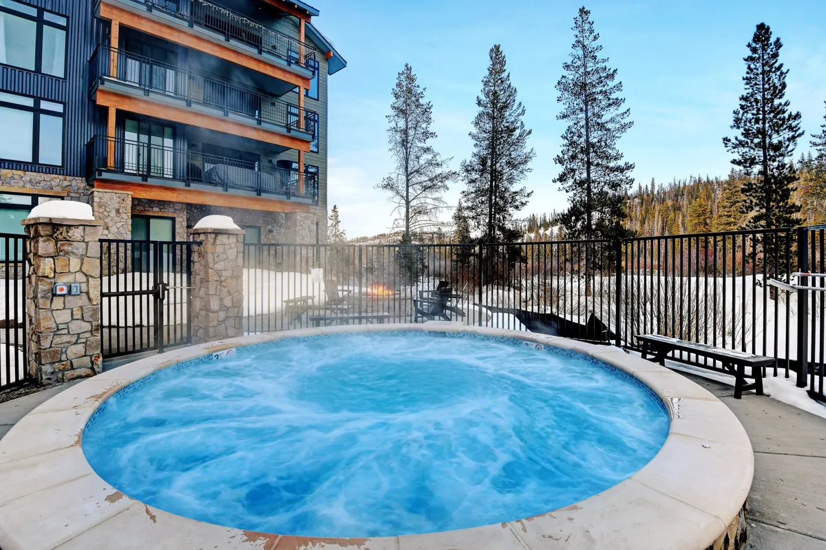 An inviting outdoor hot tub surrounded by trees, with the building facade in the background. Steam rises from the water, suggesting post-adventure relaxation in a tranquil, social setting.