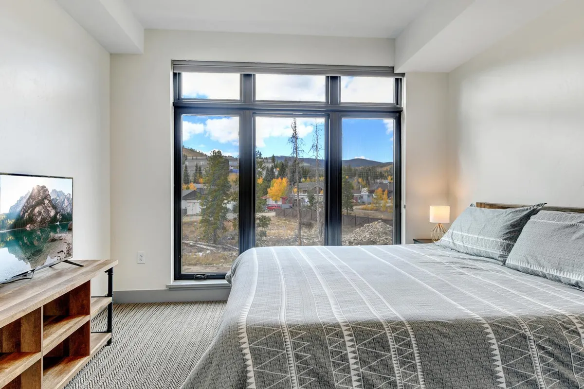A bright and cozy bedroom with large windows framing stunning mountain views, inviting guests to imagine waking up to spectacular scenery in any season. The comfortable bed and warm ambiance suggest both relaxation and connection to the outdoors.