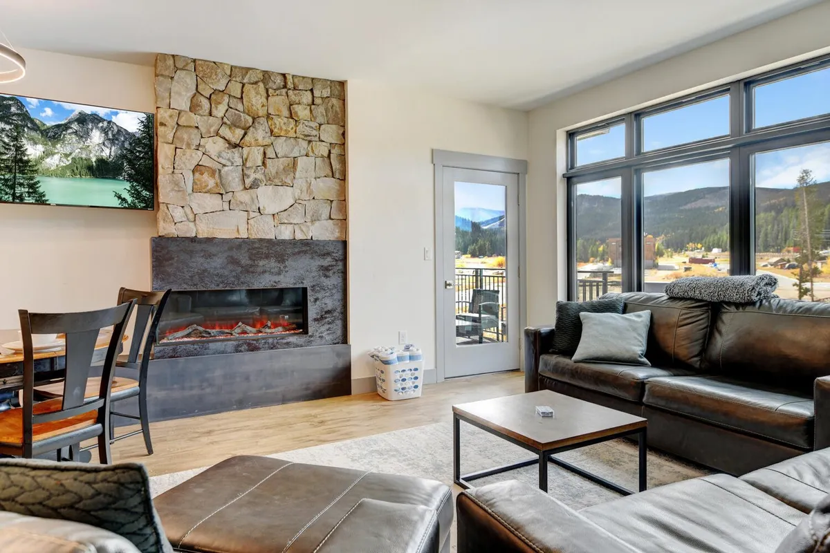 This image showcases a comfortable living area with a prominent fireplace, large windows framing mountain views, a stone accent wall, a television, and a plush sofa arranged for socializing. The room is airy, inviting, and designed for relaxation or entertaining guests.