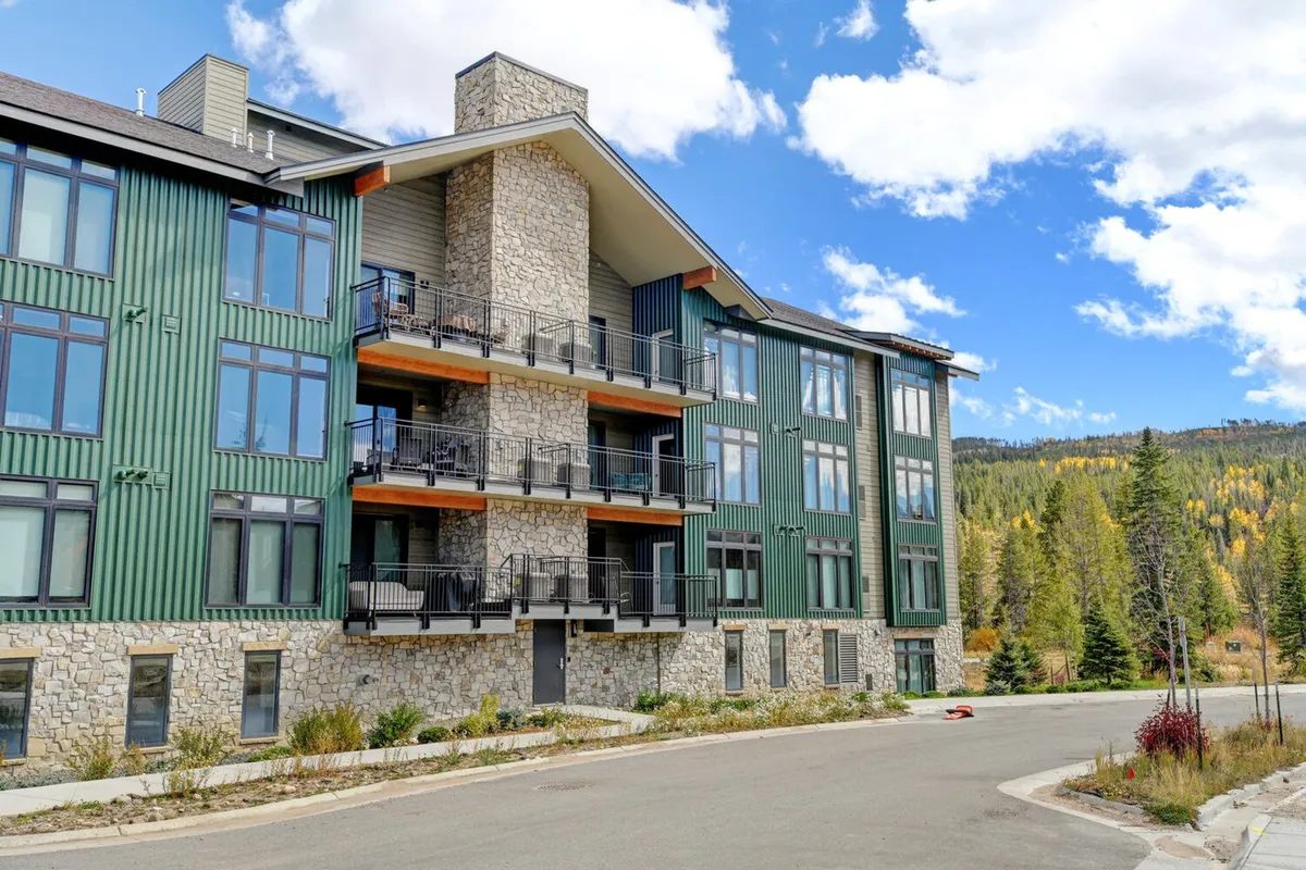 A welcoming exterior shot of the luxury condo building, featuring a stone facade, expansive windows, and private balconies overlooking the surrounding scenery. The image evokes a sense of exclusivity and modern mountain living, with hints of outdoor relaxation spaces perfect for enjoying the views.