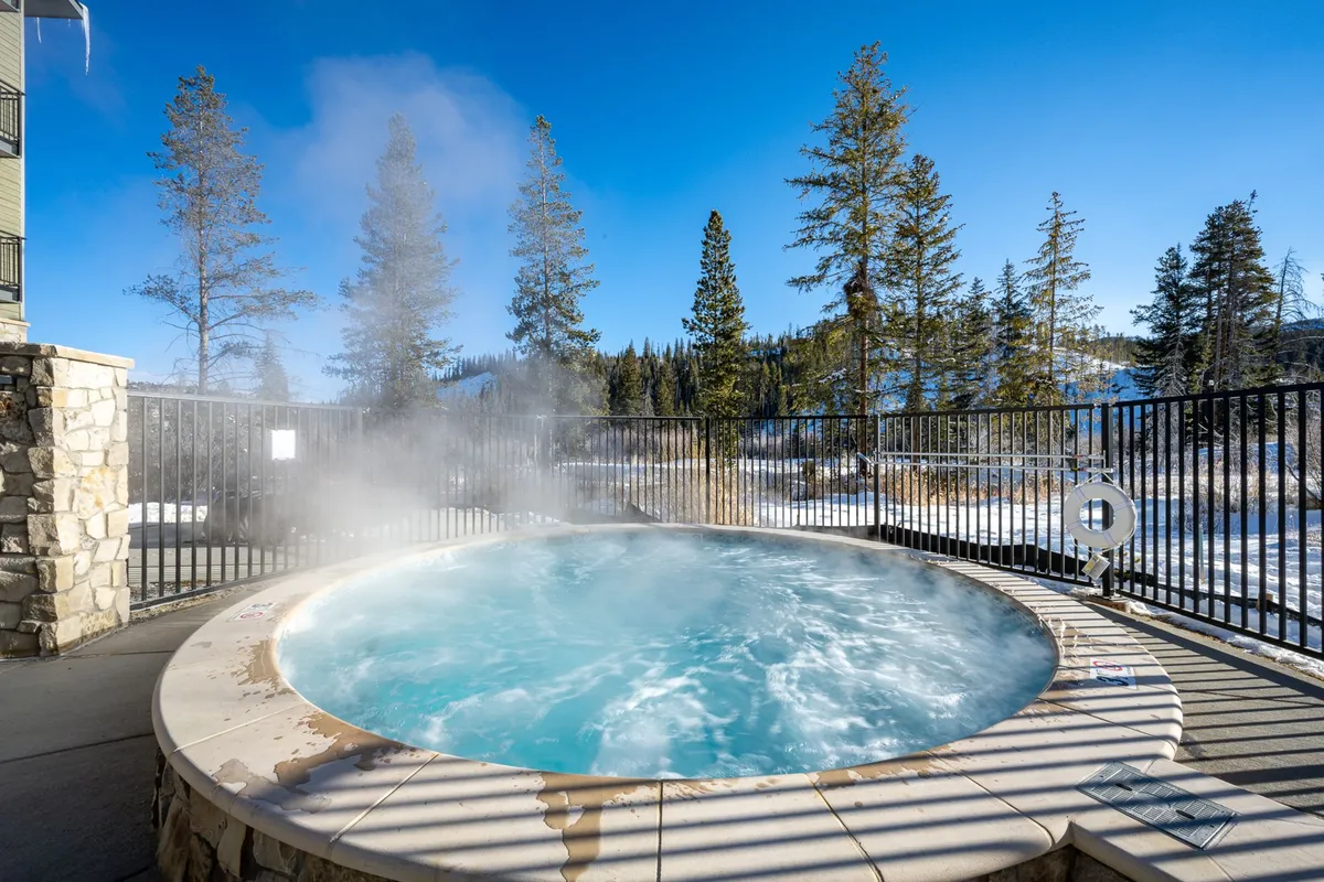 An inviting outdoor hot tub surrounded by trees, with steam rising into the crisp air. People are either relaxing in the tub or gathering nearby, creating a tranquil apres-adventure atmosphere.