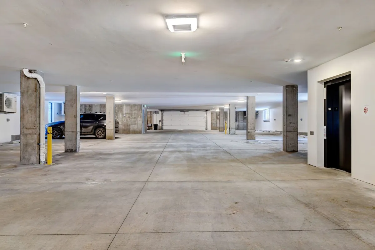 A view of the underground parking area featuring an elevator entrance, parked car, and concrete pillars. The well-lit, modern space suggests safety and convenience, with easy access to building amenities.
