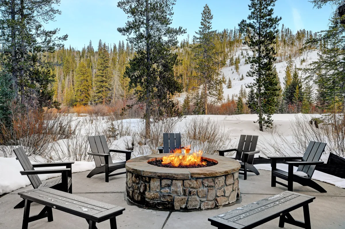 A cozy outdoor fire pit area with surrounding seating, set against a snowy alpine landscape. People gather around the fire, sharing stories and enjoying warm drinks under the open sky.