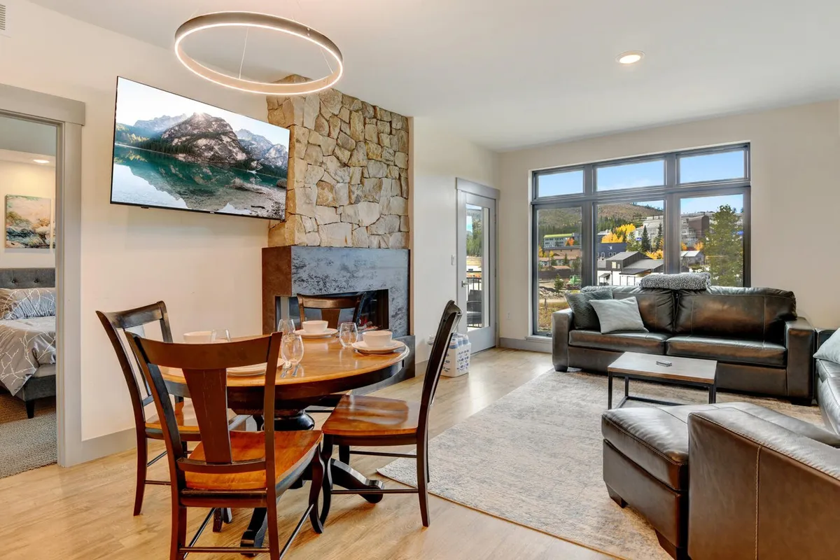 This cozy interior scene shows a dining table adjacent to a sofa, a television strategically placed to face the living area, and large windows offering mountain views. The overall atmosphere is welcoming and intimate, ideal for shared meals and relaxation.